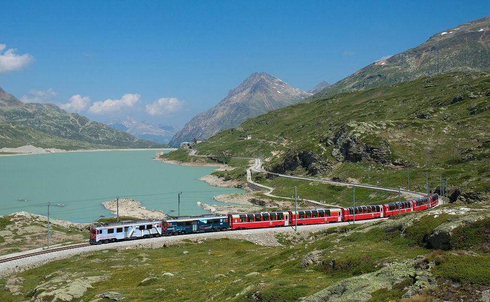 A red train crossing a mountain landscape beside a turquoise lake